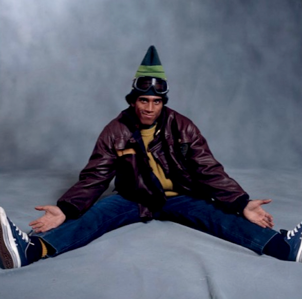 Frosty Freeze seated on a studio floor, legs spread, palms open, in a pointed knit hat and goggles
